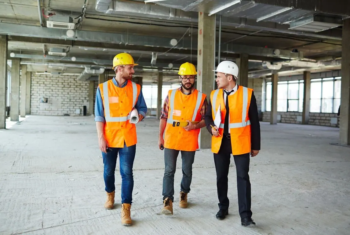 Three construction workers in hard hats and vests walking inside a building under construction.