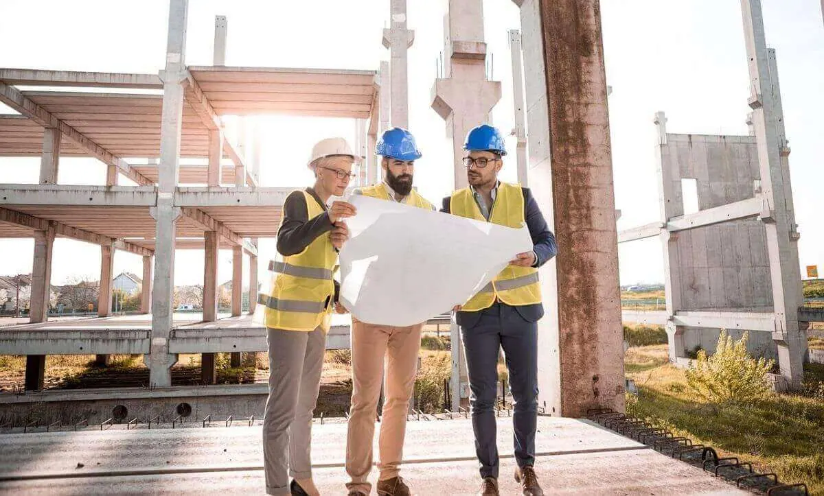 Three construction workers in hard hats examine blueprints at a building site.