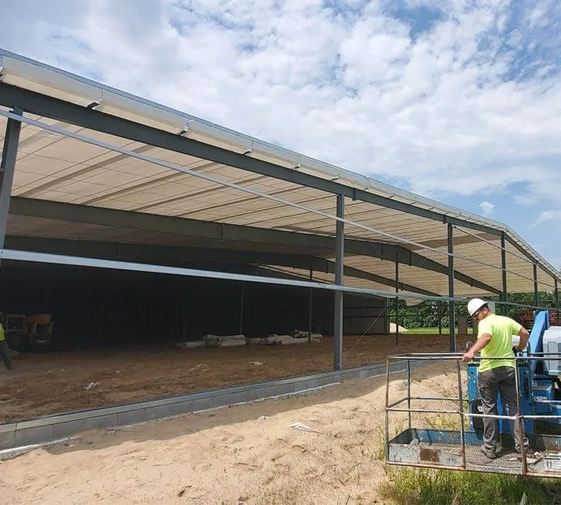 Construction worker on a lift near a large steel-framed building with a white roof and dirt floor.