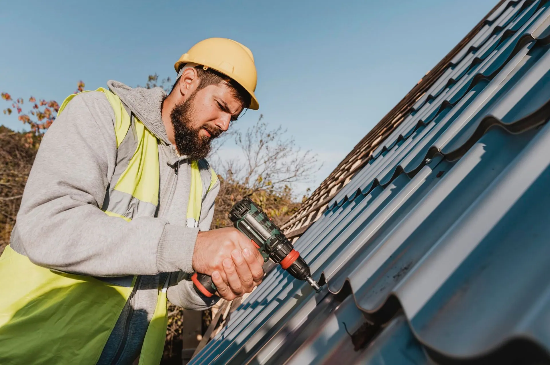 Roofer in yellow hard hat, installing metal roofing with a power drill outdoors