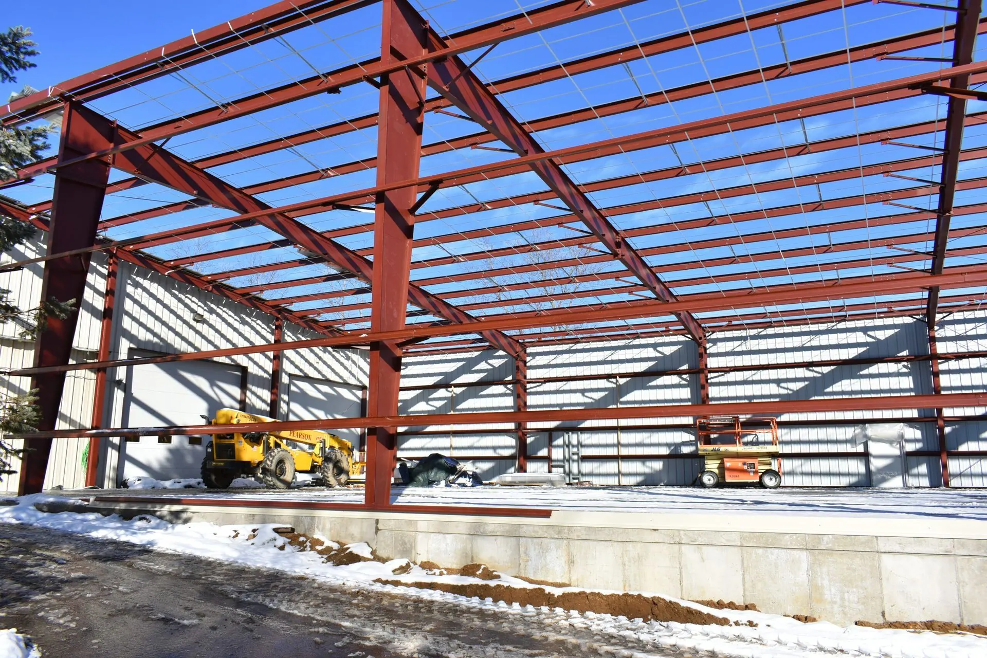 Steel frame of a building under construction, red-brown beams against a blue sky, snow on the ground.